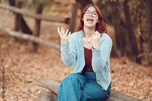 Red-haired girl screams in surprise and emotionally gesticulates with her hands. Bench in an autumn park, yellow foliage.