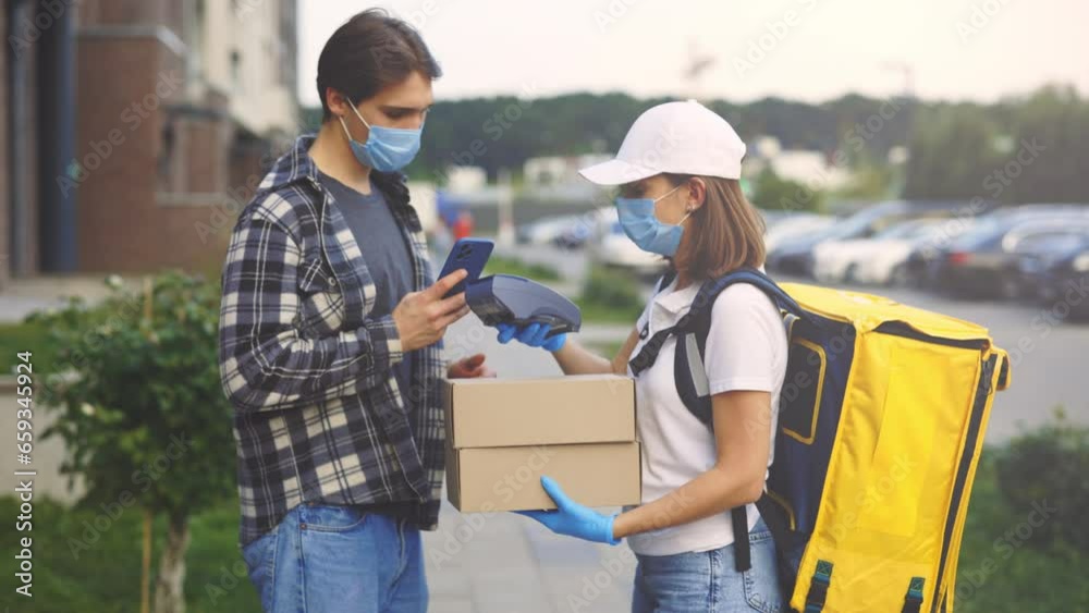Courier Lady Giving Parcel to Handsome Man Customer Wearing Protective Mask. Home Delivery While Pandemic. The Client Pays for the Package Online in Quarantine. Contactless NFC Terminal Payment Courier Lady Giving Parcel to Handsome Man Customer Wearing Protective Mask. Home Delivery While Pandemic. The Client Pays for the Package Online in Quarantine. Contactless NFC Terminal Payment