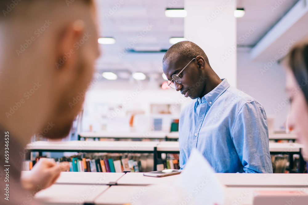 Interested ethnic student reading textbook while standing beside bookshelf in university