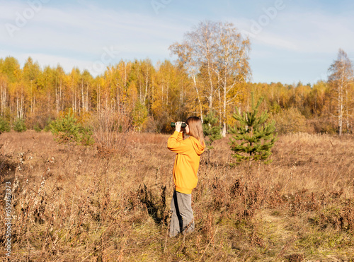 Young woman ecologist or wildlife manager in yellow hoodie with binoculars watching birds in the autumn forest Birdwatching, zoology, ecology Research, observation of animals Ornithology © Lena_viridis