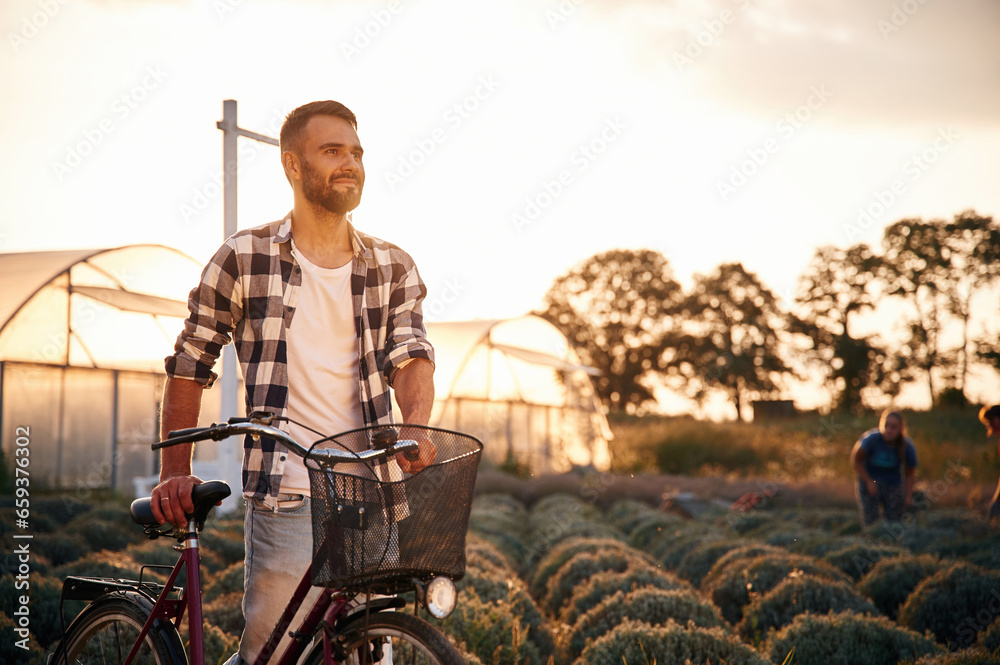 Against sunlight. Handsome man in casual clothes is with bicycle on the agricultural field near greenhouse