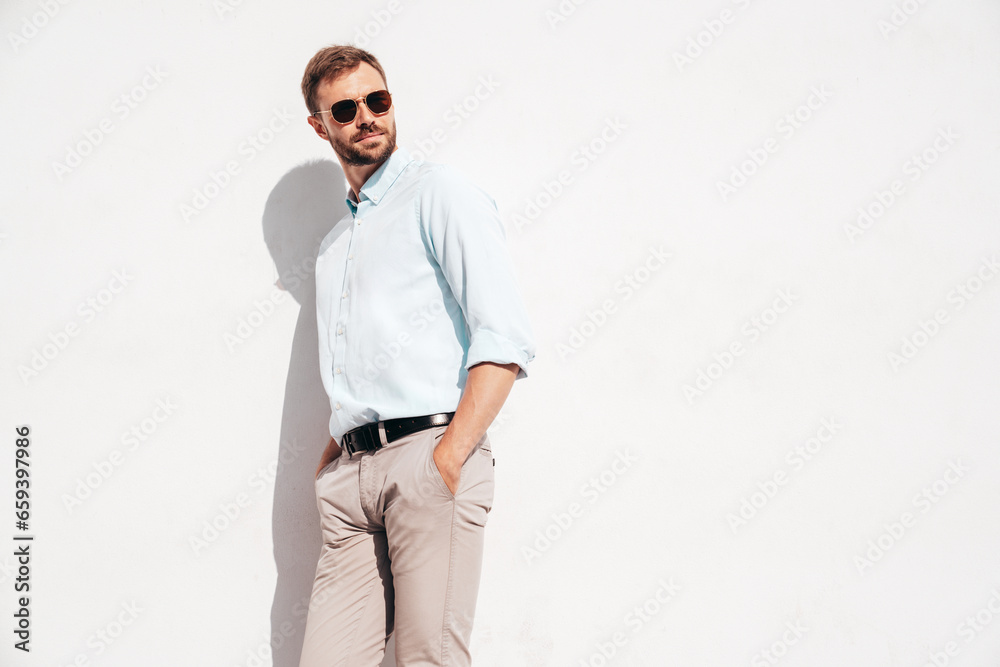 Handsome stylish hipster lambersexual model. Modern man dressed in white shirt and trousers. Fashion male posing in the street in sunglasses near white wall Handsome stylish hipster lambersexual model. Modern man dressed in white shirt and trousers. Fashion male posing in the street in sunglasses near white wall