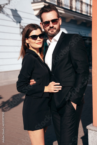  Beautiful fashion woman and her handsome elegant boyfriend in black suit. Sexy brunette model in jacket. Fashionable smiling couple posing in street. Brutal man and female outdoors. In sunglasses
