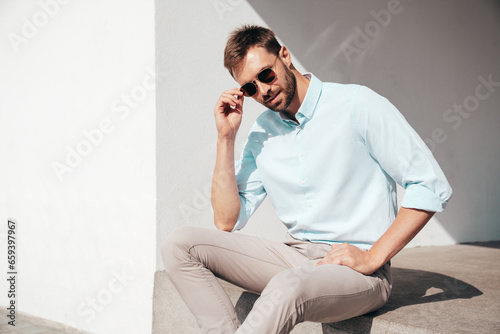 Handsome confident stylish hipster lambersexual model. Modern man dressed in white shirt and trousers. Fashion male posing in the street urban background in sunglasses. Outdoors, sitting