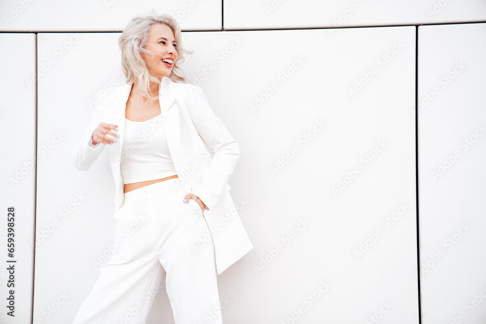 Young beautiful blond woman wearing nice trendy white suit jacket. Smiling model posing in the street at sunny day. Fashionable female outdoors. Cheerful and happy. posing near wall Young beautiful blond woman wearing nice trendy white suit jacket. Smiling model posing in the street at sunny day. Fashionable female outdoors. Cheerful and happy. posing near wall