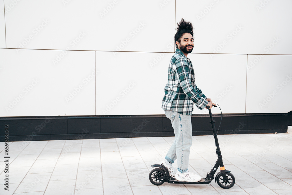 Trendy smiling bearded man in casual clothes riding electric scooter in urban background. Handsome model posing in the street near wall. Hipster guy with curly hairstyle