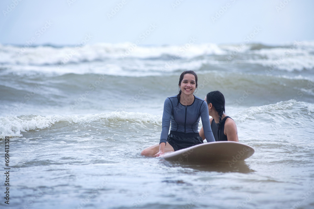 Pretty teen girl smiling and sitting on surfboard on the sea, learn to surf with a female instructor, women and outdoor sports