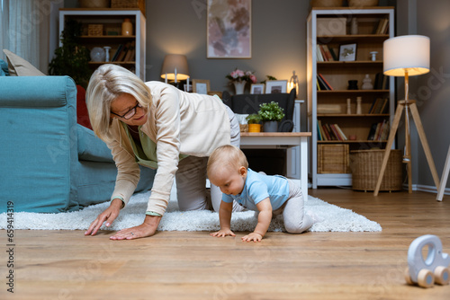 Grandmother of single mother taking care of her young baby, playing on the apartment floor. Senior woman babysitting her grandson or granddaughter while child parent are at work or date night.