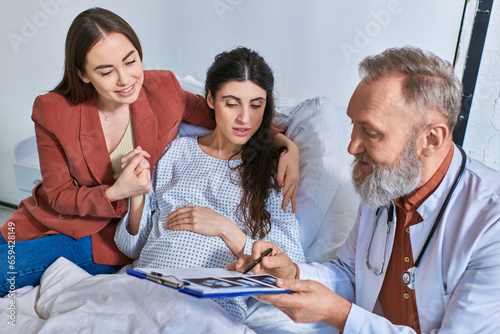 cheerful grey bearded doctor showing ultrasound to lesbian couple, in vitro fertilization concept