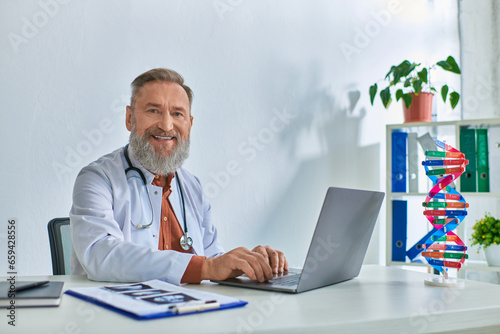 happy grey bearded doctor with ultrasound on table working on laptop and smiling sincerely at camera