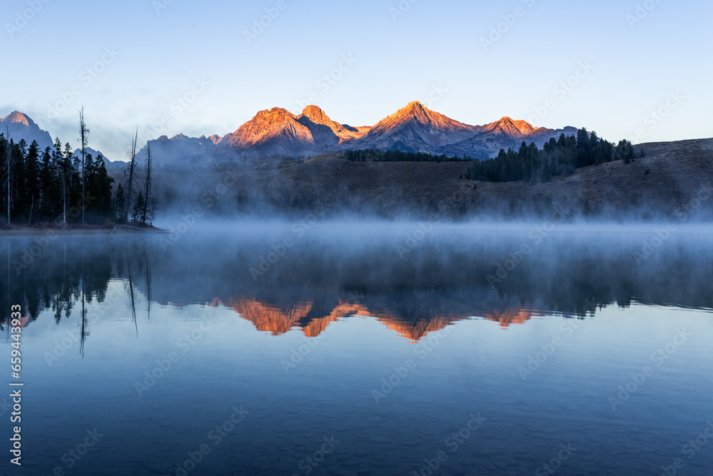 Redfish lake in Idaho in dawn. Calm water covered by mist. Mountains range is reflecting in lake water - obrazy, fototapety, plakaty 