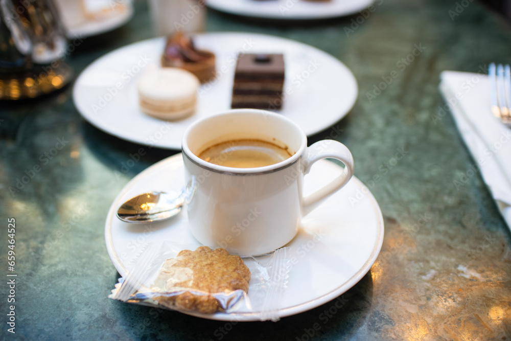 A cup of black espresso coffee with spoon and cookie, a plate of assorted dessert such as opéra pastry, macaron and mini tart in background on white table with spoon. A cup of black espresso coffee with spoon and cookie, a plate of assorted dessert such as opéra pastry, macaron and mini tart in background on white table with spoon.