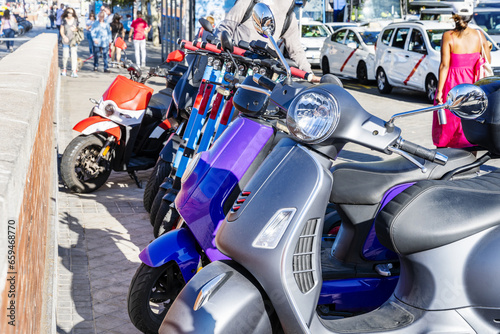 Several motorcycles and mopeds of all kinds and a row of blue electric scooters standing on a sidewalk in a European city
