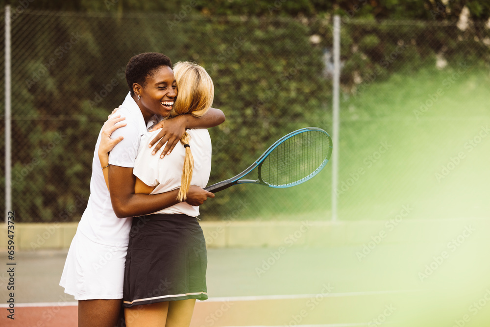 Portrait of two happy young beautiful women with tennis clothes and rackets in a tennis court ready to play a game. Two friends sharing a morning of sportive activity playing a tennis match. - obrazy, fototapety, plakaty 