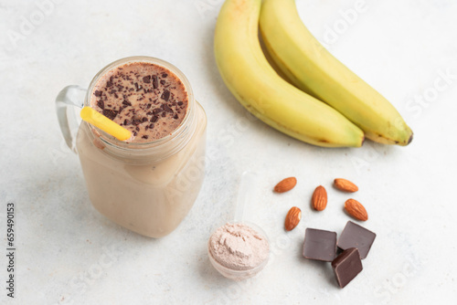Glass jar of protein milkshake drink or smoothie and whey protein powder in measuring spoon, bananas, chocolate cubes, almond nuts on white background. sport nutrition, bodybuilding food supplements