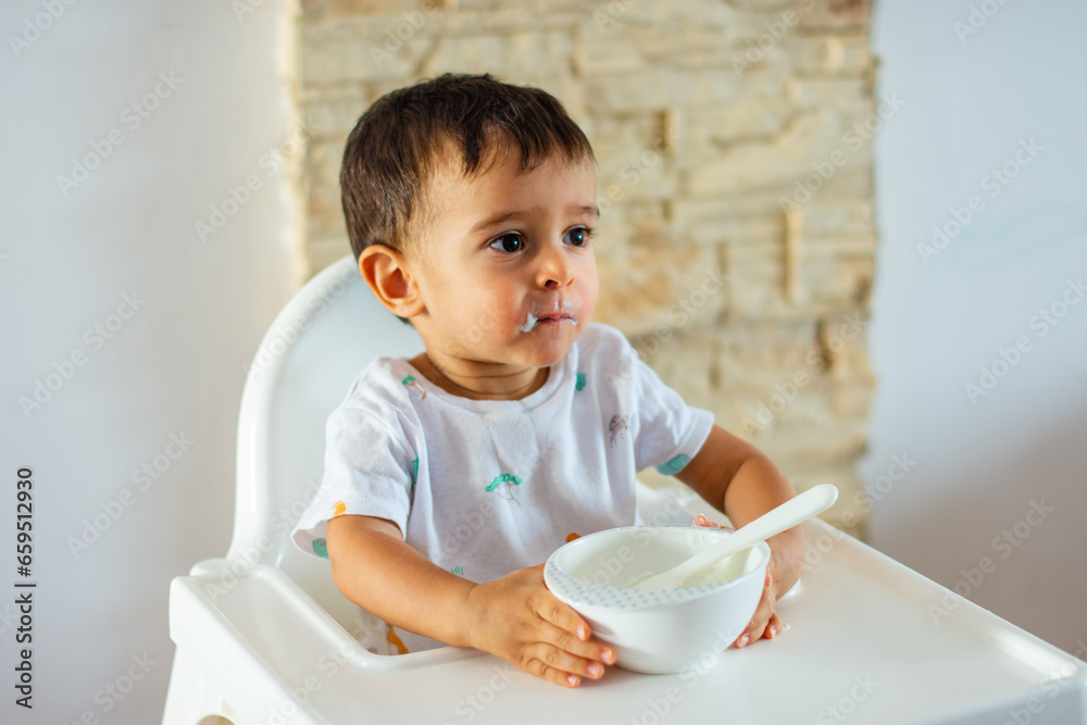 Toddler doesn't want to eat anymore holding his plate while his face is stained with yogurt Toddler doesn't want to eat anymore holding his plate while his face is stained with yogurt