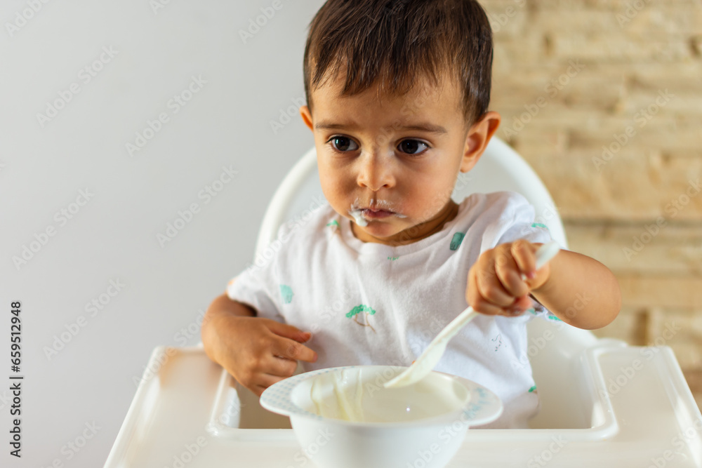 Cute little Caucasian boy tasting and enjoying Greek yogurt alone with his spoon while sitting in a baby chair Cute little Caucasian boy tasting and enjoying Greek yogurt alone with his spoon while sitting in a baby chair