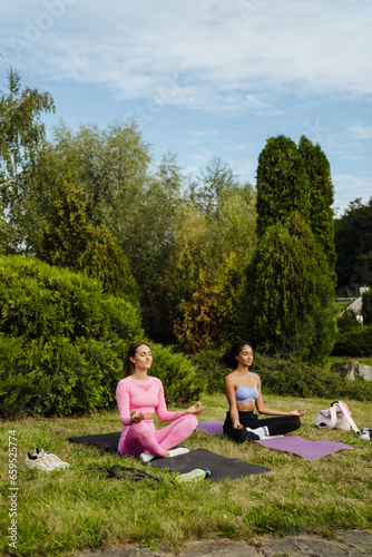 Two young women sitting on yoga mats in lotus pose and meditating in green park
