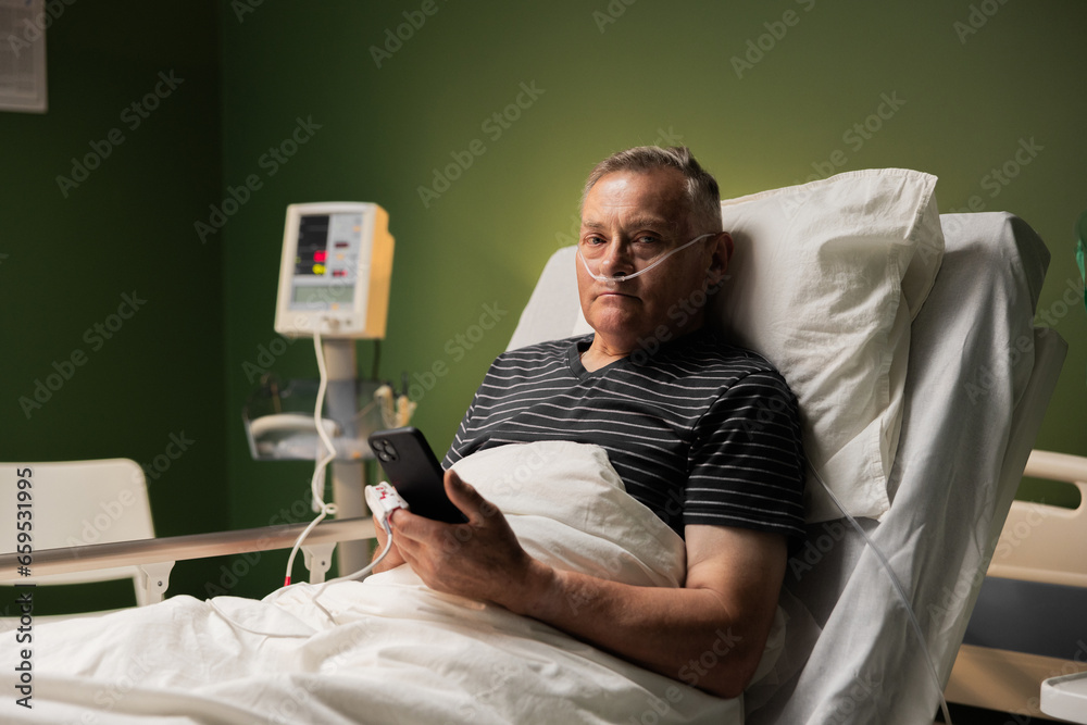 Elderly man in a hospital bed, holding cellphone, and wearing an oxygen tube and finger monitor, hospitalized due to pneumonia Elderly man in a hospital bed, holding cellphone, and wearing an oxygen tube and finger monitor, hospitalized due to pneumonia