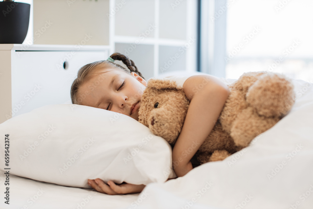 Cute caucasian girl sleeping sweet on white soft pillows under blanket and hugging her teddy bear friend. Pretty kid with two braids regaining energy during disease at home. Cute caucasian girl sleeping sweet on white soft pillows under blanket and hugging her teddy bear friend. Pretty kid with two braids regaining energy during disease at home.