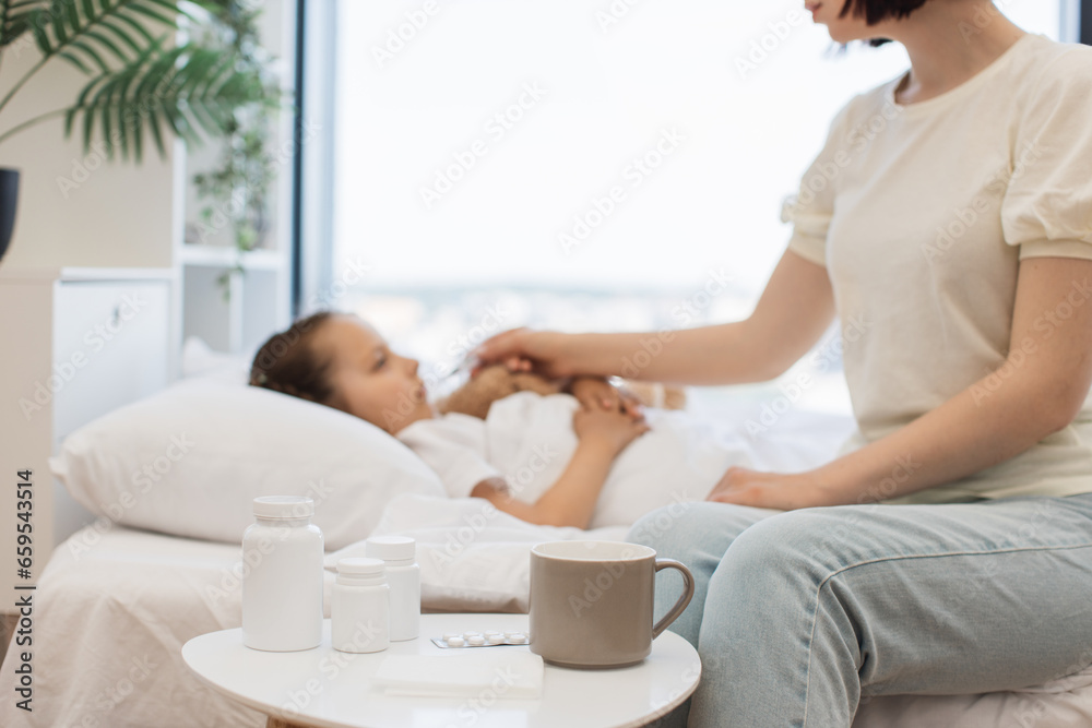 Fototapeta premium Focus on bedside table with cup of hot beverage, paper tissues and medical pills standing near white bed. Blurred background of caring mother measuring daughter's fever with digital thermometer.