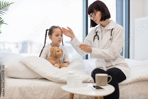Young girl patient sitting in white bed under watchful care of doctor conducting thorough examination and checking temperature. Cute child hugging toy and preparing to taking medicine.