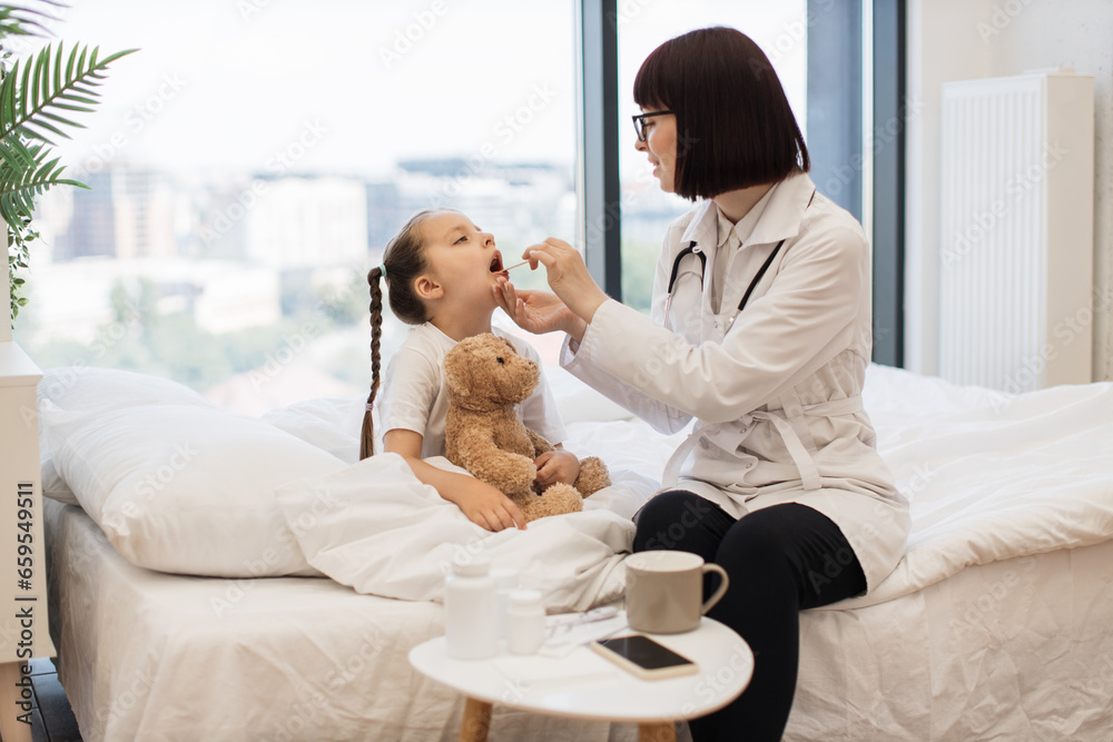 Young girl sitting on white bed while caring female doctor examining her throat in cozy bedroom. Tired caucasian child feeling pain and need treatment under supervision of physician. Young girl sitting on white bed while caring female doctor examining her throat in cozy bedroom. Tired caucasian child feeling pain and need treatment under supervision of physician.