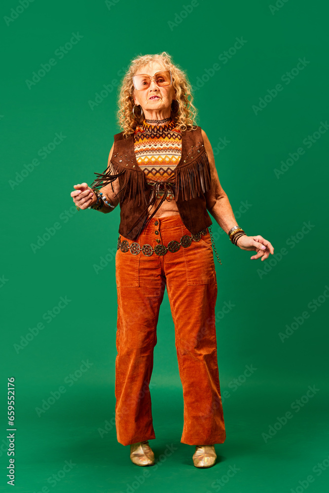 Hippie style. Extravagant senior woman in stylish cloths and accessories standing over green studio background. Concept of human emotions, fashion, elderly people, lifestyle, creativity. Ad