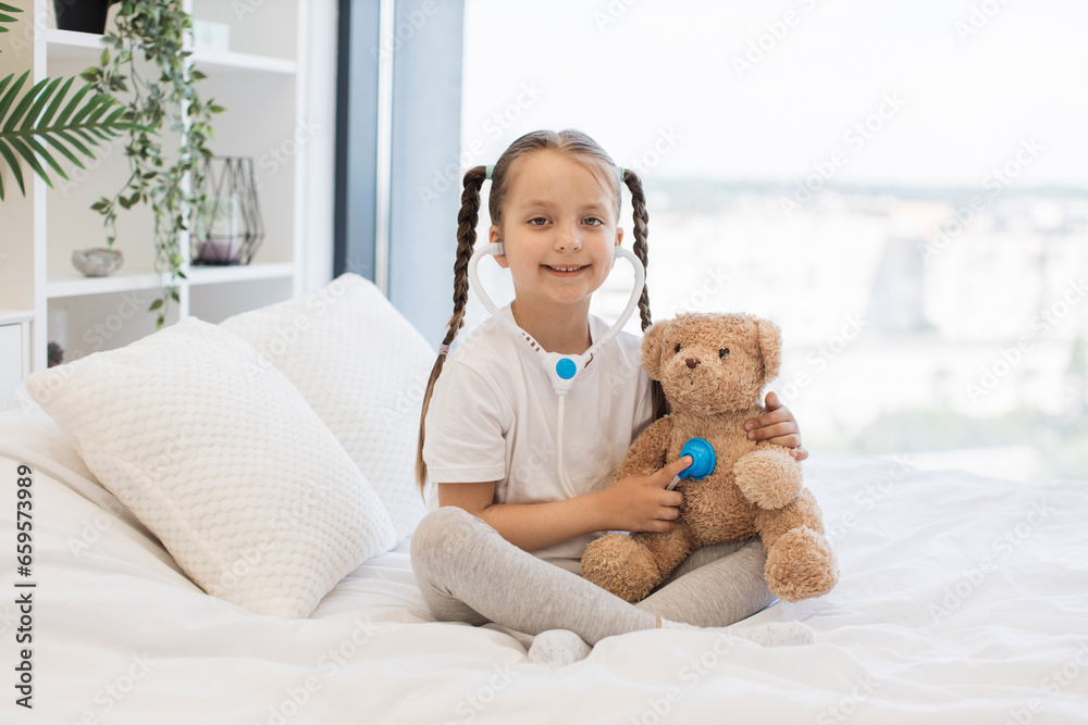 Portrait of beautiful caucasian girl relaxing on white cozy bed with brown soft teddy bear in well-lighted room. Charming child using toy stethoscope to examining fluffy ill patient. Portrait of beautiful caucasian girl relaxing on white cozy bed with brown soft teddy bear in well-lighted room. Charming child using toy stethoscope to examining fluffy ill patient.