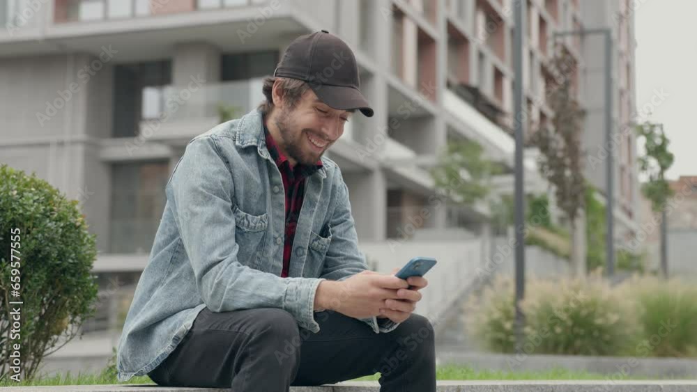Young Man In Casual Dressed Using Phone While Sitting on Street Against Background of City Building Young Man In Casual Dressed Using Phone While Sitting on Street Against Background of City Building