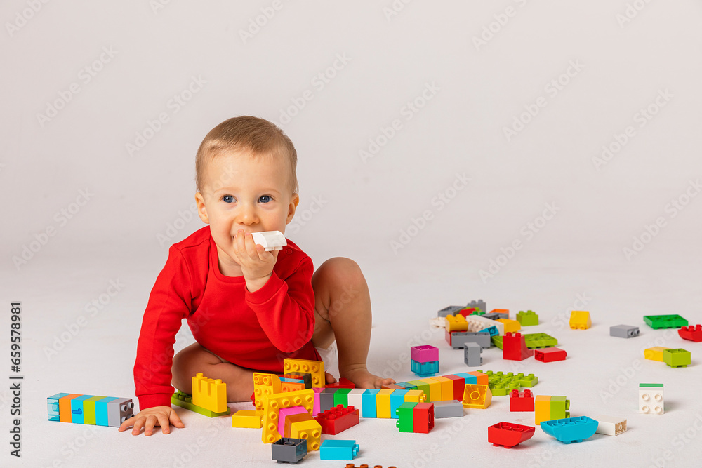 Cute baby boy in red bodysuit playing with a multicolored plastic constructor on a white background isolate, child development concept, space for text Cute baby boy in red bodysuit playing with a multicolored plastic constructor on a white background isolate, child development concept, space for text