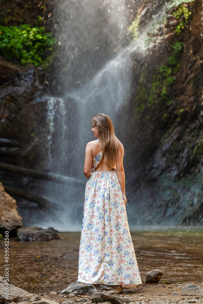 Woman in dress, tropical waterfall backdrop. Nature adventure, water flow, travel concept. Forest, scenic view. Summer tropical vacation, outdoors adventure, lush jungle.