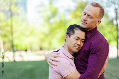 Portrait of middle aged gay couple, homosexual men hugging with closed eyes in park, outdoors
