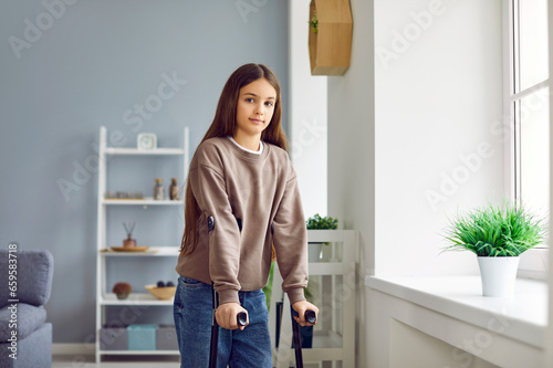 Portrait of child with leg injury. Little kid with crutches. Little girl with orthopedic crutches standing in living room at home and looking at camera. Rehabilitation concept