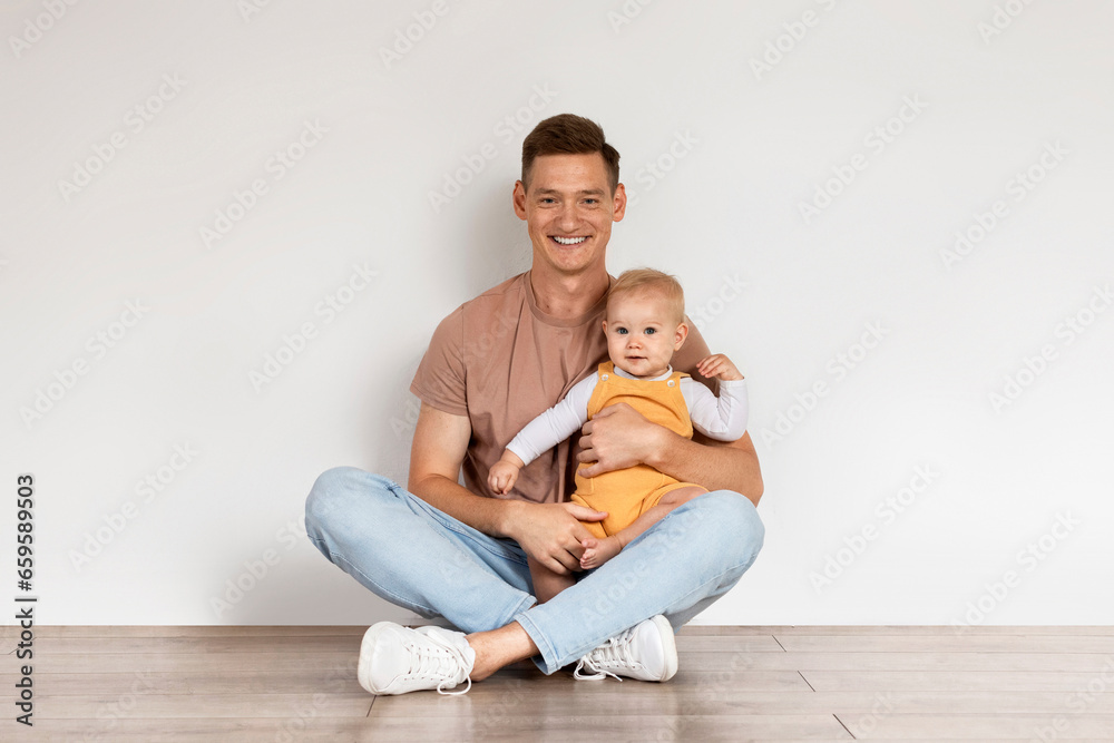 Happy Young Dad With Infant Son Sitting On Floor Near White Wall Happy Young Dad With Infant Son Sitting On Floor Near White Wall