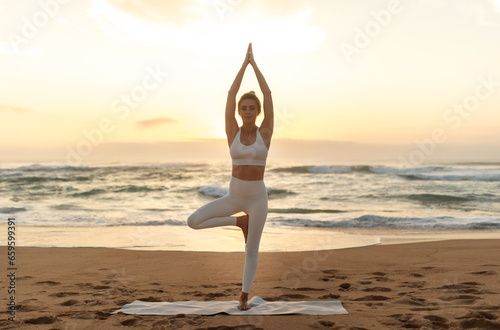 Fit woman standing in tree pose on fitness mat, lady practicing yoga outdoors on sea beach, full length shot