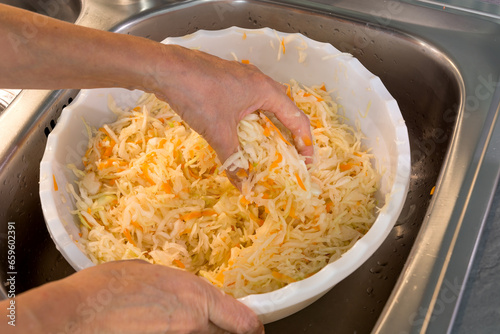 Preparing homemade sauerkraut for the winter. Women's hands knead and mix finely chopped cabbage and carrots with salt in a plastic basin in a metal sink in the kitchen. Real photo. photo
