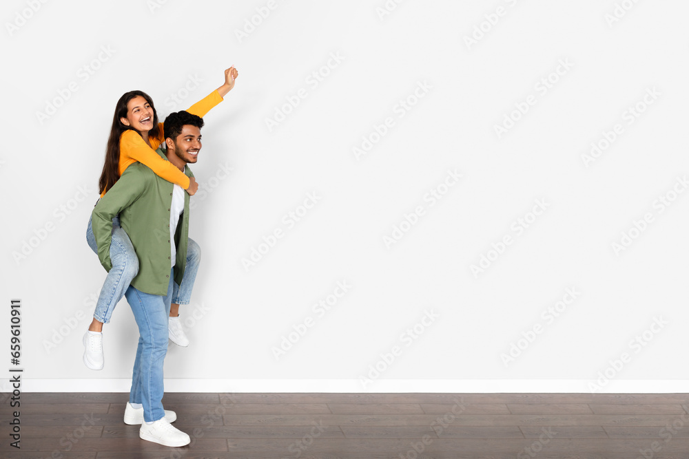 Joyful indian man piggybacking wife, woman raising arm, free space