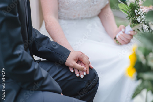 Ein Brautpaar sitzt nebeneinander, hält sich liebevoll an den Händen, die Braut trägt ein weißes Kleid und hält einen Blumenstrauß, der Bräutigam einen dunklen Anzug.