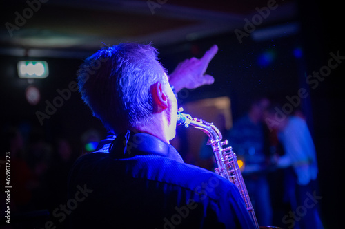 Ein Musiker spielt Saxophon auf einer Bühne in einer Bar oder bei einer Party, stimmungsvolles blaues Licht, der Musiker zeigt mit dem Arm nach oben, unscharfer Hintergrund mit Publikum.