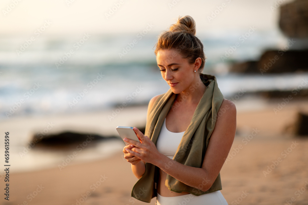 Sporty woman with smartphone resting after outdoors training, browsing fitness app, standing on sea beach, copy space