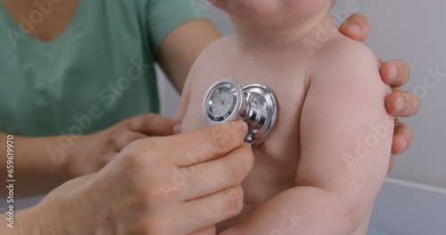 Handsome child sits at doctor checkup and puts pacifier in mouth. Pediatrician uses stethoscope to listen heartbeat and lungs of little kid. Mother with cute baby in hospital. Close up. Slow motion.