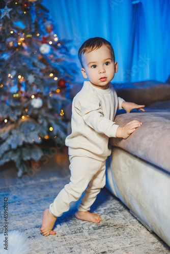 Little handsome one-year-old boy dressed in a beige suit on the background of a Christmas tree in a home environment. Children's emotions before celebrating the New Year holiday
