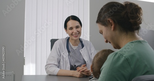 Female pediatrician consults patient with cute little child on appointment in hospital. Woman sits on chair at physician checkup. Friendly doctor works in modern medical center. Close up. Back view.