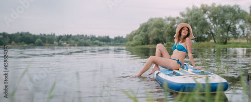 Canvas Print A young woman with an open swimsuit swims on a SUP board on a picturesque lake