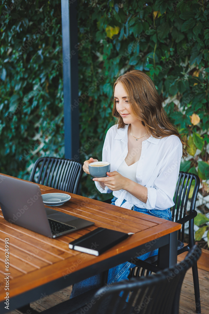 Young woman in casual clothes drinking coffee and talking on the phone, working on a green terrace at a laptop, looking at the screen and working on a project remotely from work in a cafe Young woman in casual clothes drinking coffee and talking on the phone, working on a green terrace at a laptop, looking at the screen and working on a project remotely from work in a cafe