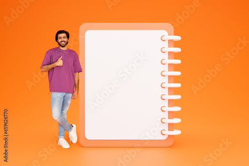 Smiling young indian guy in casual show thumb up with hands near list with free space, approve work planning