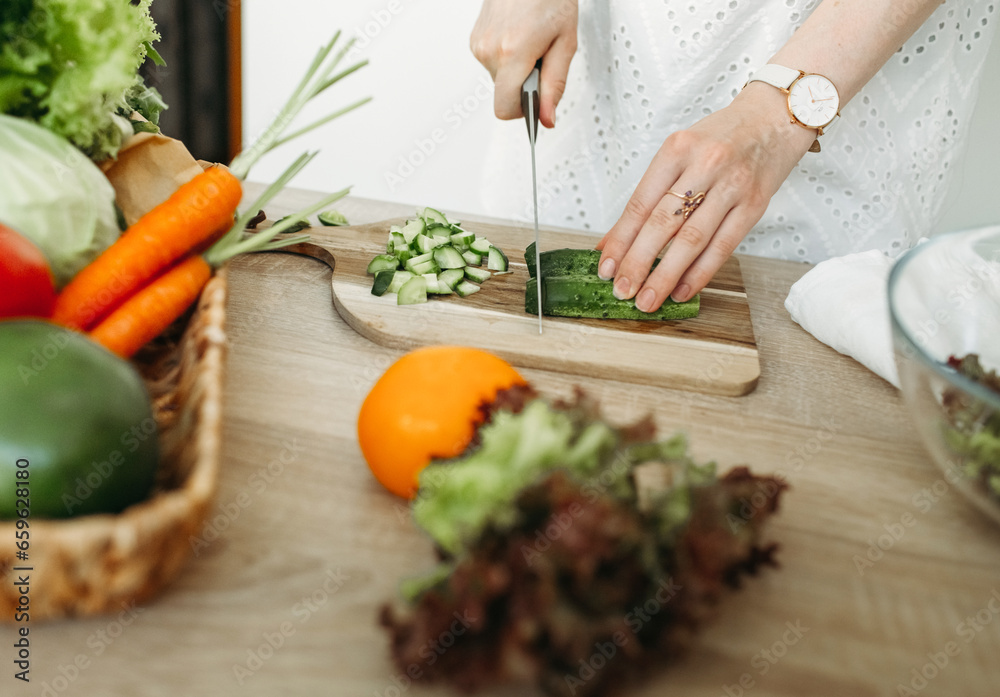 A woman is cutting a cucumber, vegetables lie next to her on the kitchen table. A woman is cutting a cucumber, vegetables lie next to her on the kitchen table.