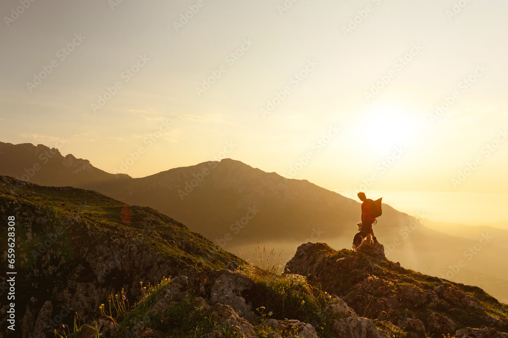 Mountaineer man equipped with a backpack contemplates the high mountain landscape at sunset while practicing hiking with his dog. Outdoor sports and adventure