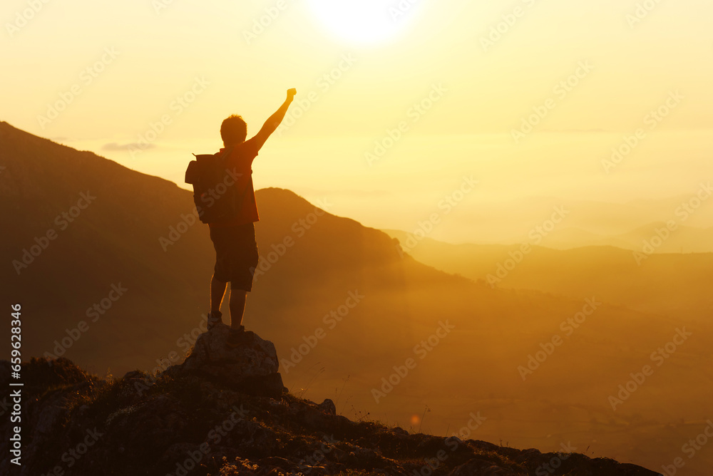 Silhouette of a mountaineer man with his fist raised in victory celebrating his ascension to a mountain peak at sunset. sport and outdoor adventure.